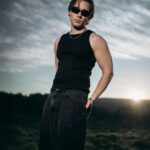 A confident man in sunglasses posing outdoors at sunset, wearing a black tank top and jeans.