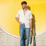 Casually dressed man stands against a vibrant yellow wall holding a bag under sunlight.