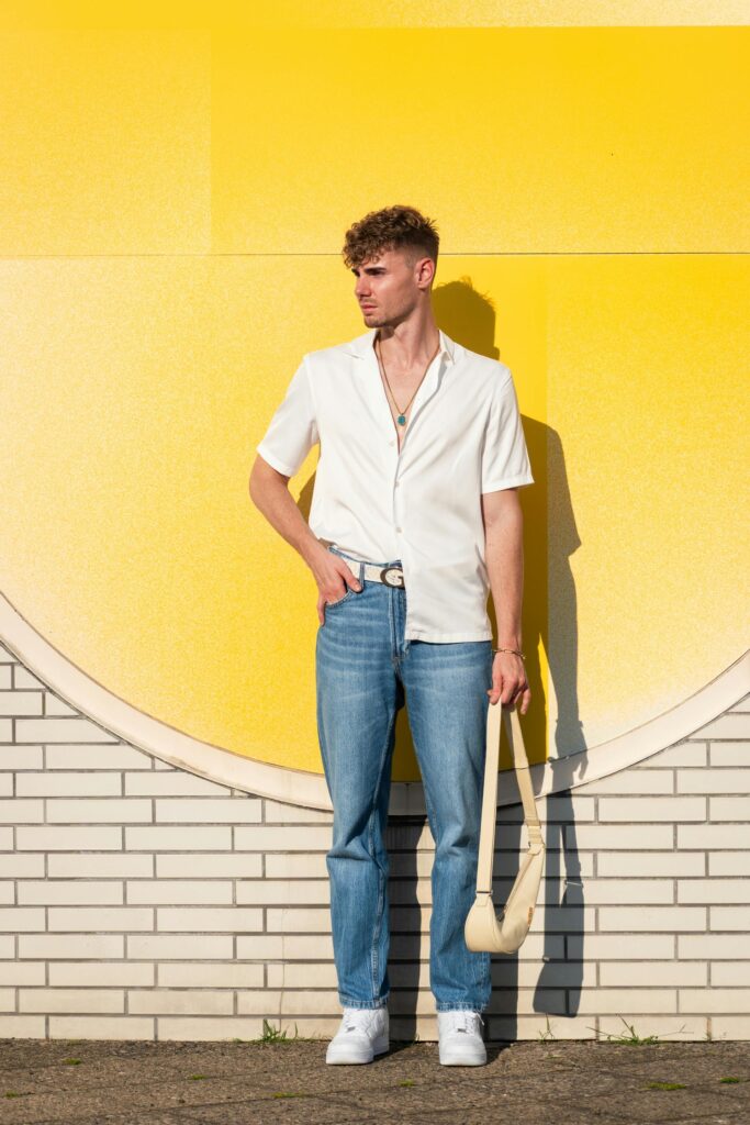 Casually dressed man stands against a vibrant yellow wall holding a bag under sunlight.
